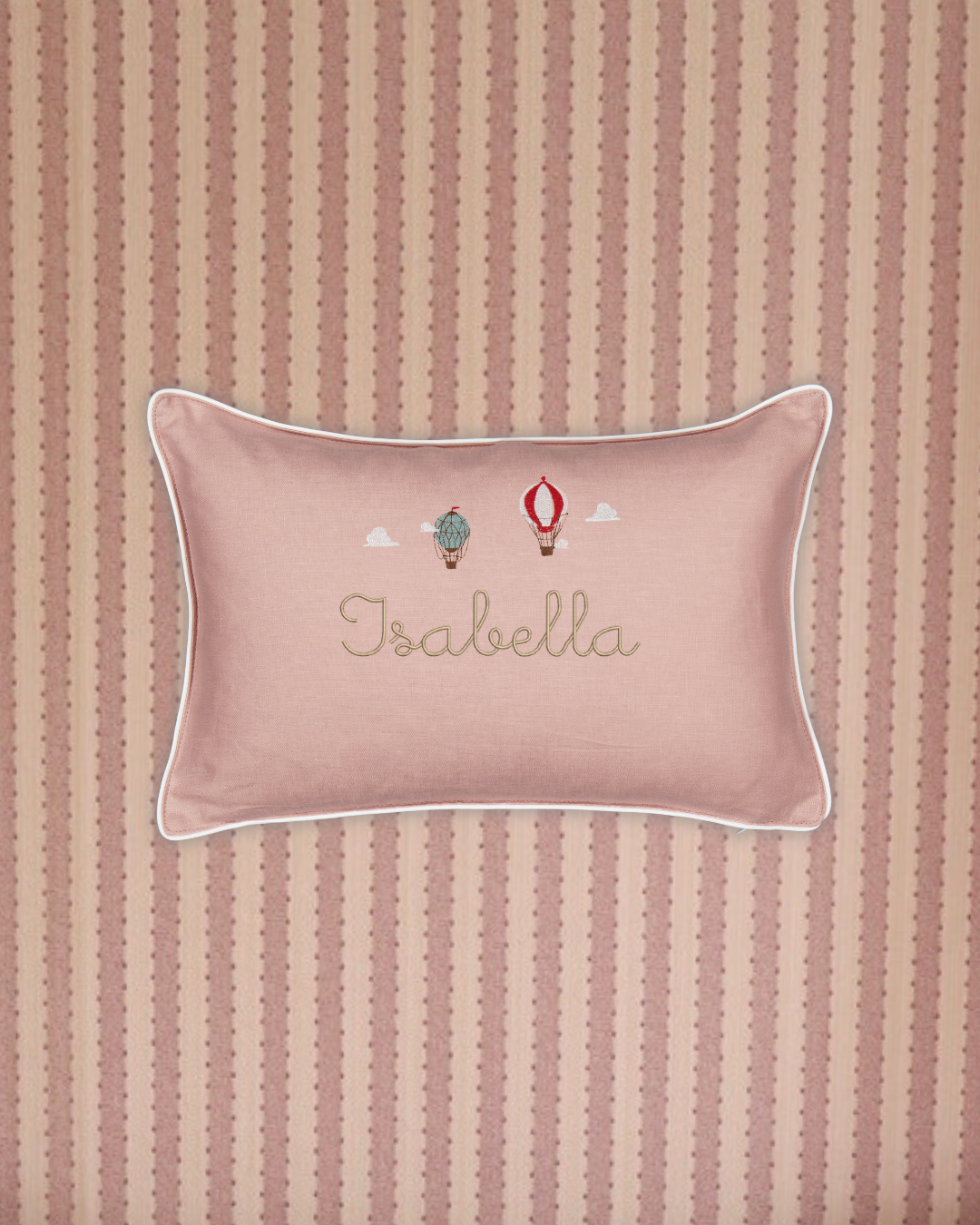 Pink linen cushion with white piping, embroidered hot air balloons and personalized name "Isabella" on striped fabric background