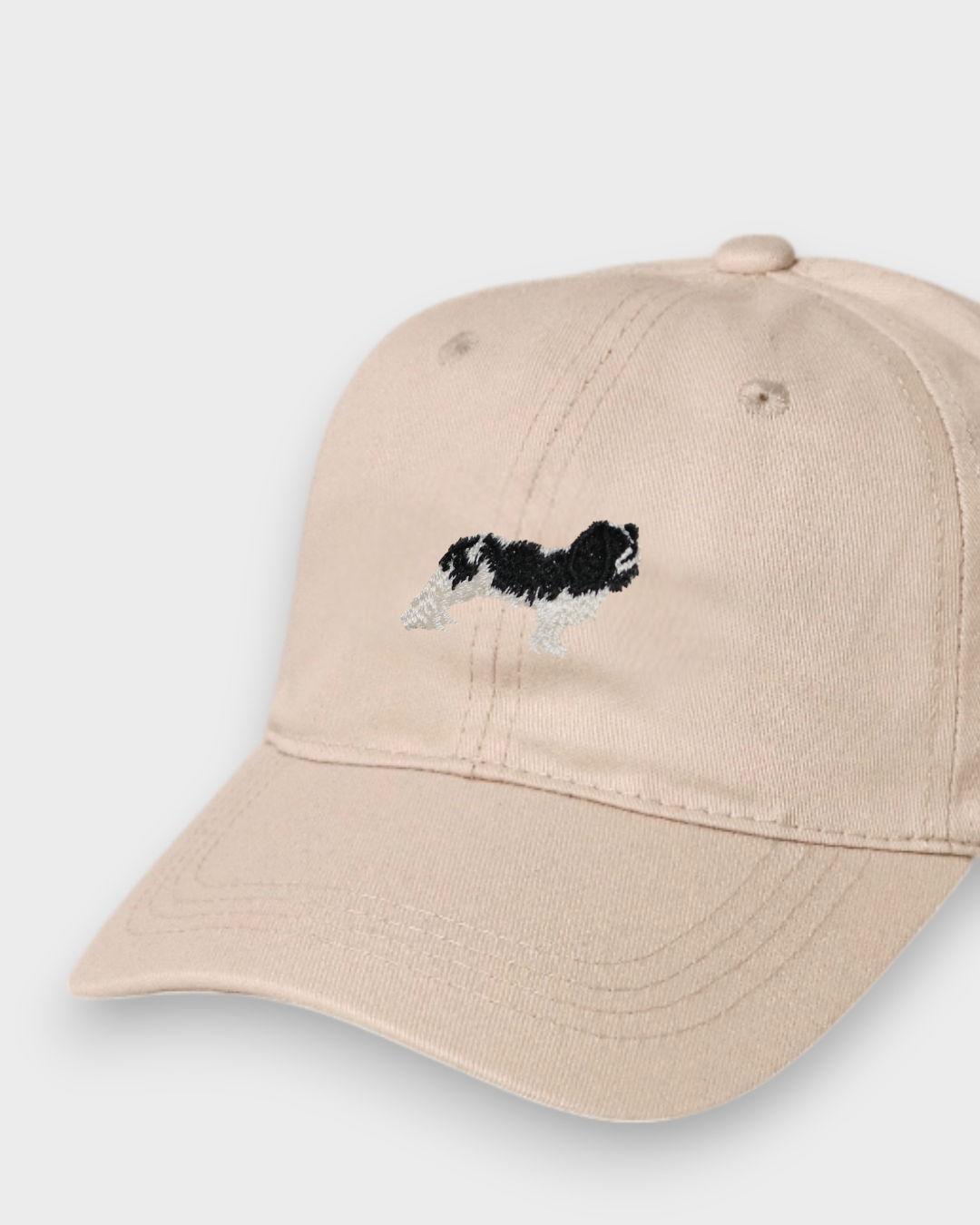 Beige baseball cap with embroidered black and white King Charles Spaniel design, personalized embroidery option on back, made from 100% washed cotton.
