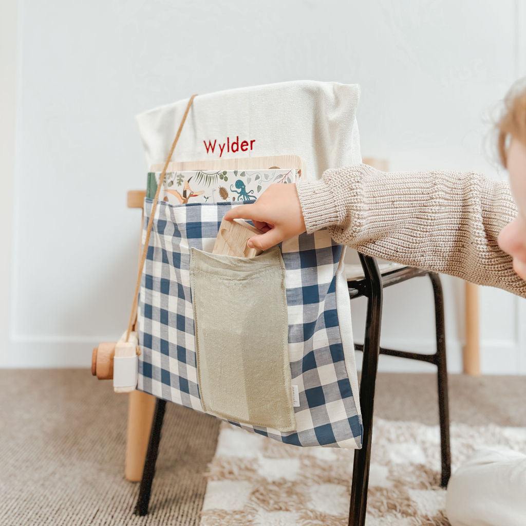 Personalised blue gingham chair bag on a child's chair, featuring embroidered name and front pocket for organization.