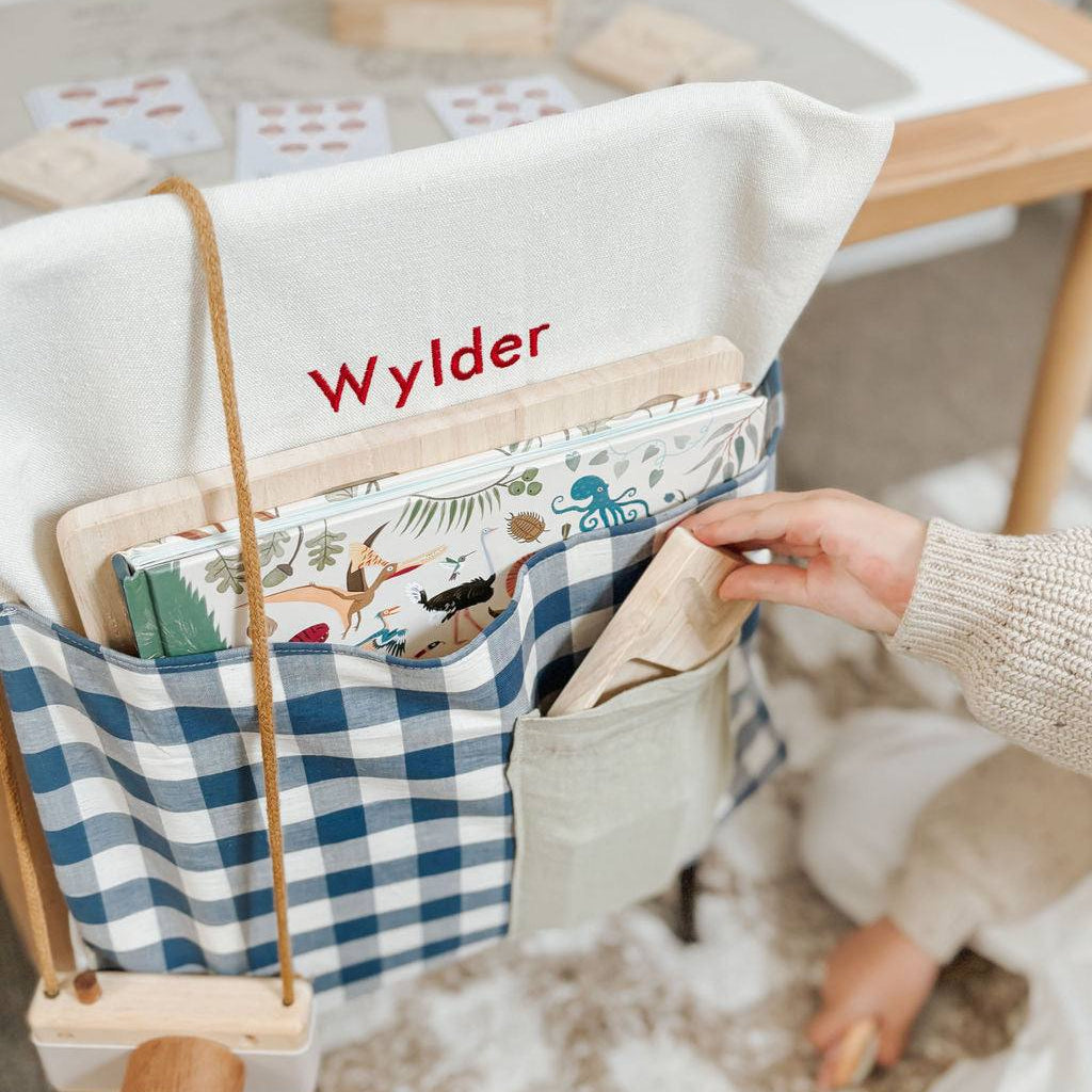 Blue gingham personalised chair bag on a child's chair, embroidered with "Wylder," holding books and stationery.