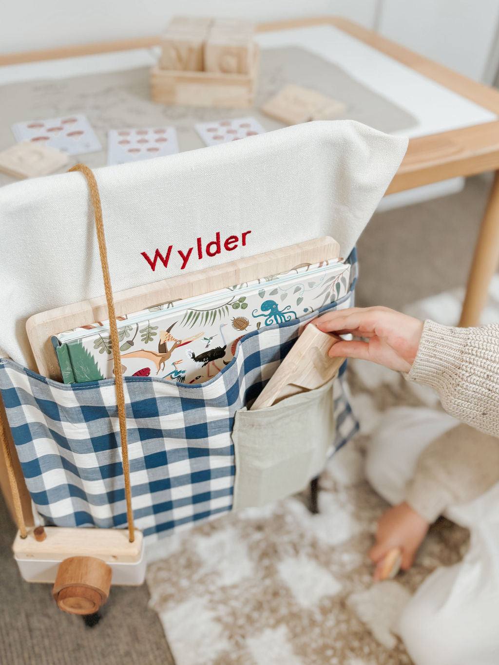 Blue gingham personalised chair bag on a child's chair, embroidered with "Wylder," holding books and stationery.