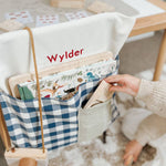 Blue gingham personalised chair bag on a child's chair, embroidered with "Wylder," holding books and stationery.