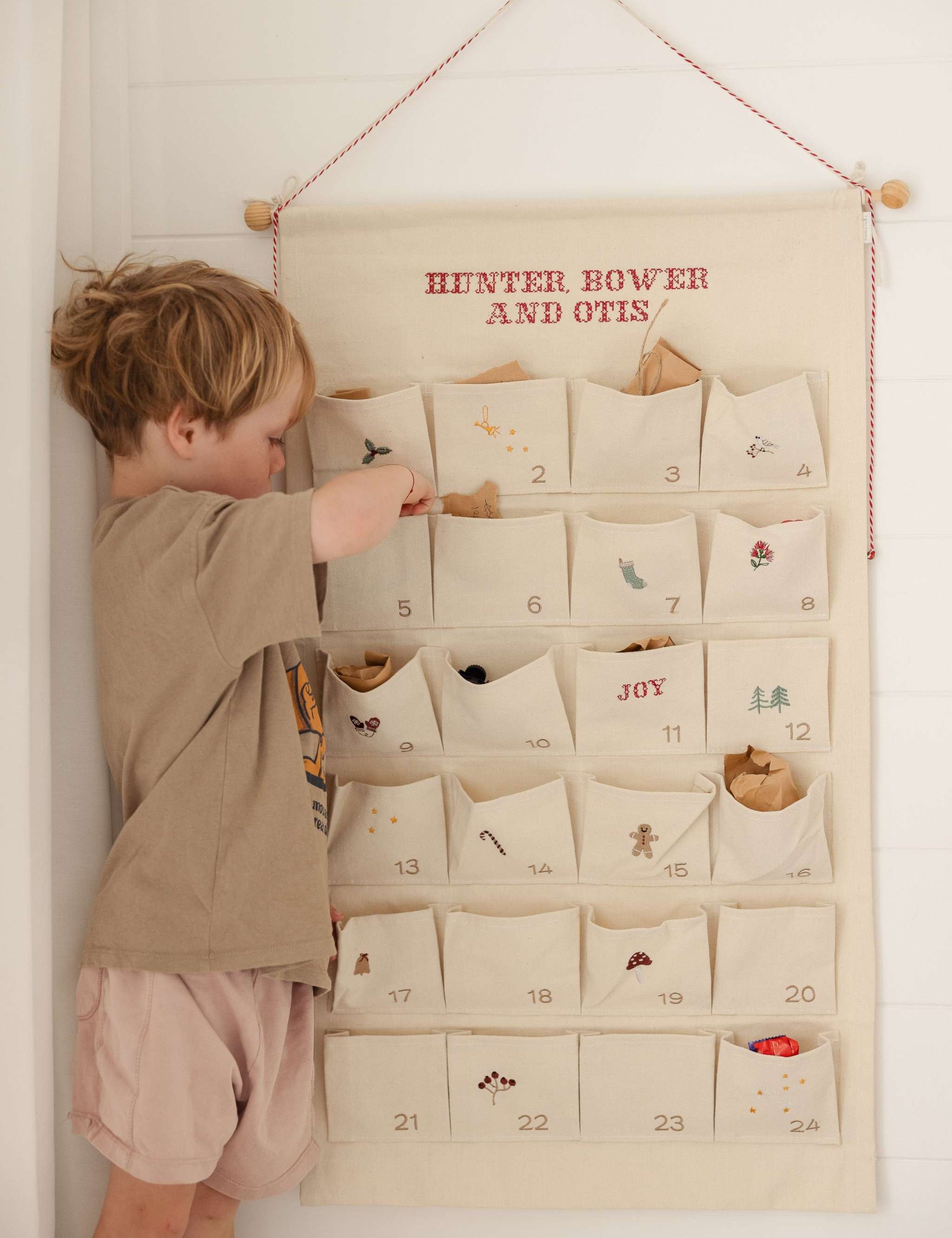 Child interacting with ivory cotton canvas embroidered advent calendar featuring personalized red text and 24 numbered pockets with whimsical designs and tiny bells on hanging string